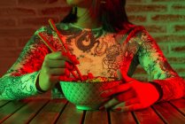 Неэмоциональная молодая азиатка с иероглифами на лице в стильной шляпе, поедающая вкусную лапшу, сидя в темной комнате напротив кирпичной стены — стоковое фото