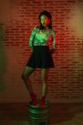 Corpo inteiro confiante bonita Asiática fêmea em mini-saia e chapéu elegante de pé graciosamente em barril de metal e olhando para a câmera durante sessão de fotos contra parede de tijolo em estúdio — Fotografia de Stock