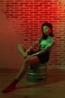 Plein corps confiant jolie Asiatique femelle en mini jupe et chapeau élégant assis gracieusement sur le canon en métal et en regardant la caméra pendant la séance photo contre le mur de briques en studio — Photo de stock
