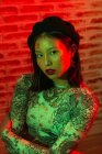 Неэмоциональная молодая азиатка в стильной одежде с иероглифами на лице, спокойно смотрящая в камеру на кирпичную стену в студии — стоковое фото