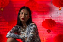Jovem mulher asiática sem emoção no desgaste elegante com hieróglifos pintados no rosto olhando para a câmera calmamente contra a parede de tijolo no estúdio — Fotografia de Stock