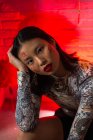 Привлекательная азиатская женщина с раскрашенными иероглифами на лице в стильном наряде, уверенно стоящая и нежно трогательно смотрящая в камеру в современной студии — стоковое фото