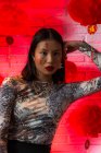 Attraente donna asiatica alla moda con geroglifici dipinti sul viso che indossa un vestito elegante seduto con fiducia e toccando teneramente il viso mentre guarda la fotocamera in studio moderno — Foto stock