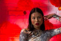 Attraktive, trendige Asiatin mit aufgemalten Hieroglyphen im Gesicht in stilvollem Outfit, die selbstbewusst sitzt und ihr Gesicht zärtlich berührt, während sie in ein modernes Studio blickt — Stockfoto