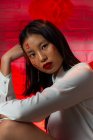 Attraktive, trendige Asiatin mit aufgemalten Hieroglyphen im Gesicht in stilvollem Outfit, die selbstbewusst steht und ihr Gesicht zärtlich berührt, während sie in ein modernes Studio blickt — Stockfoto