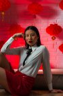 Attraente donna asiatica sicura di sé con geroglifici dipinti sul viso indossando minigonna rossa seduta sul pavimento e guardando la fotocamera durante la sessione fotografica contro il muro di mattoni in studio — Foto stock