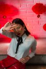 Atractiva mujer asiática confiada con jeroglíficos pintados en la cara con mini falda roja sentada en el suelo y mirando a la cámara durante la sesión de fotos contra la pared de ladrillo en el estudio - foto de stock