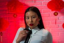 Atractiva mujer asiática confiada con jeroglíficos pintados en la cara con mini falda roja sentada en el suelo y mirando a la cámara durante la sesión de fotos contra la pared de ladrillo en el estudio - foto de stock