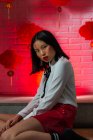Приваблива приваблива азіатська жінка з ієрогліфами намальовані на обличчі у червоній міні-спідниці сидячи на підлозі і дивлячись на камеру під час фотосесії проти цегляної стіни в студії. — стокове фото