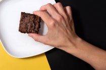 Mulher mão pegar pedaço de brownie fresco no fundo preto e amarelo — Fotografia de Stock
