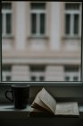 Libro di testo con cerchi da bevanda a pagina vicino tazza contro finestra e edificio in sera — Foto stock