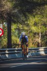 Full body of young sportsman in activewear and helmet riding bicycle on asphalt road amidst lush green trees on sunny day — Stock Photo
