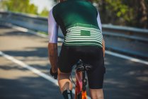 Back view of young sportsman in activewear and helmet riding bicycle on asphalt road amidst lush green trees on sunny day — Stock Photo