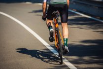Вид сзади на молодого спортсмена в спортивной одежде и шлеме на велосипеде на асфальтированной дороге среди пышных зеленых деревьев в солнечный день — стоковое фото