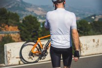 Вид сзади на неузнаваемого молодого велосипедиста в защитном шлеме и спортивной одежде, идущего рядом с велосипедом, припаркованным на асфальтированной дороге против голубого неба — стоковое фото