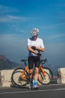 Полное тело молодого велосипедиста в защитном шлеме и спортивной одежде, идущего рядом с велосипедом, припаркованным на асфальтированной дороге против голубого неба — стоковое фото