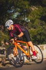 Ganzkörper-Seitenansicht eines jungen Sportlers in Aktivkleidung und Helm, der an sonnigen Tagen auf kurvenreicher Asphaltstraße inmitten sattgrüner Bäume Fahrrad fährt — Stockfoto