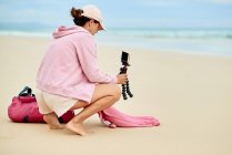 Side view of full body of female travel blogger putting cellphone on tripod in sand for shooting video for social media — Stock Photo