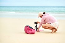 Side view full body of female travel blogger putting cellphone on tripod in sand for shooting video for social media — Stock Photo