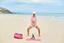 Full body of happy active female traveler squads during training on sandy beach and recording video on mobile phone — Stock Photo