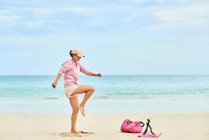 Vue latérale du corps complet du voyageur féminin actif soulevant le genou pendant l'entraînement sur la plage de sable et enregistrement vidéo sur téléphone portable — Photo de stock