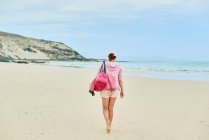 Voltar ver corpo inteiro de turista feminino anônimo com saco andando descalço ao longo da costa vazia em direção ao mar ondulando — Fotografia de Stock