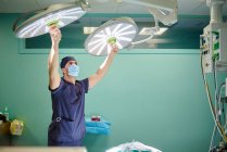 Концентрированный молодой медик в форме и маске, регулирующий хирургический свет перед операцией в современной больнице — стоковое фото