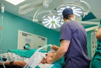 Вид со стороны анонимного анестезиолога-мужчины в медицинской форме и кепке, держащего маску анестезии на лице неузнаваемого пациента, лежащего на диване в операционной — стоковое фото