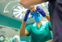 Знизу невизначеної жінки-медика допомагає врожаю чоловічому лікареві наносити латексні рукавички на руки перед операцією в сучасній клініці — стокове фото