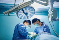 Side view of unrecognizable male doctor with assistant in medical gowns and masks performing surgery with laser in operating room — Stock Photo