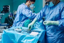 Anonimo chirurgo maschio focalizzato in maschera blu abito e lenti di ingrandimento in piedi vicino campo sterile con vari strumenti medici durante il funzionamento in clinica moderna — Foto stock