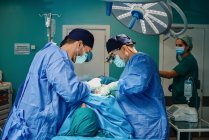 Paciente irreconocible acostado en la mesa de operaciones durante la cirugía realizado por médicos varones concentrados en batas y máscaras médicas - foto de stock