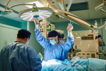 Невпізнаваний чоловічий медичний асистент, що налаштовує хірургічну лампу, стоячи біля операційного столу та лікаря в сучасній клініці — стокове фото