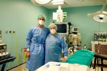 Старшие и молодые хирурги-мужчины в стерильных платьях и медицинских масках обнимаются и смотрят в камеру, стоя в операционной после работы — стоковое фото
