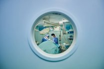 Through round window of unrecognizable doctors in uniforms and masks performing surgery in modern operating room — Foto stock