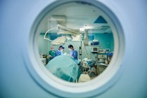 Through round window of unrecognizable doctors in uniforms and masks performing surgery in modern operating room — Stock Photo