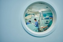 Через кругле вікно невизнаних лікарів у формі та масках, що виконують операцію в сучасній операційній кімнаті — стокове фото