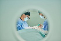 Through round window of unrecognizable doctors in uniforms and masks performing surgery in modern operating room — Foto stock
