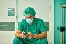 Anonymous young male doctor in green medical uniform and mask messaging on mobile phone while sitting in operating room after surgery — Stock Photo