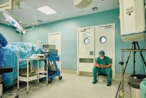 Jeune médecin anonyme homme en uniforme médical vert et masque de messagerie sur téléphone portable tout en étant assis dans la salle d'opération après la chirurgie — Photo de stock