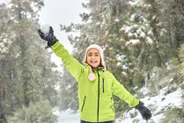 Захоплена дівчина в теплому одязі і капелюсі кидає сніжок, розважаючись у замерзлій зимовій лісі і дивлячись з щасливою посмішкою — стокове фото