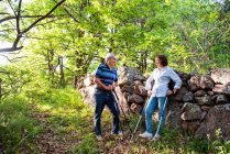 Cheerful mature couple standing on trail in green woods while having break during pole walking and looking at each other — Foto stock