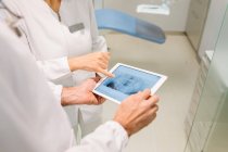 Dentistes anonymes en peignoirs médicaux examinant l'état des dents sur la radiographie sur tablette tout en travaillant ensemble dans une clinique dentaire moderne — Photo de stock