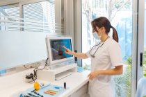 Vue de côté dentiste féminine compétente en uniforme examinant l'image de rayon X à l'écran dans la clinique dentaire moderne légère — Photo de stock