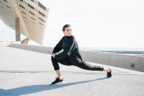 Full length determined young sportswoman in activewear stretching legs while warming up on road in suburb — Foto stock