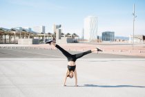 Back view full length fit determined sportswoman in tight activewear doing handstand exercise on asphalt road on sunny suburb with open legs — Fotografia de Stock