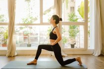 Side view of young barefoot female in sportswear standing in Anjaneyasana pose on yoga mat against window in house — Stock Photo