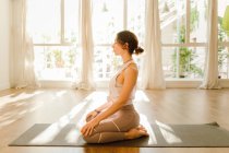 Side view of young dreamy female in sportswear practicing yoga with arms behind back while sitting with closed eyes in sunlight — Foto stock