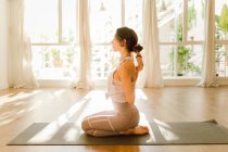 Вид збоку молодої мрійливої жінки в спортивному одязі, що практикує йогу з руками позаду, сидячи з закритими очима на сонячному світлі — стокове фото