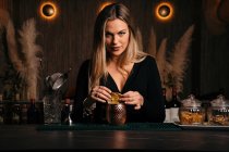 Self assured attractive young female barkeeper with long blond hair in stylish outfit decorated cocktail with lemon slices while standing at counted in stylish bar — Stock Photo